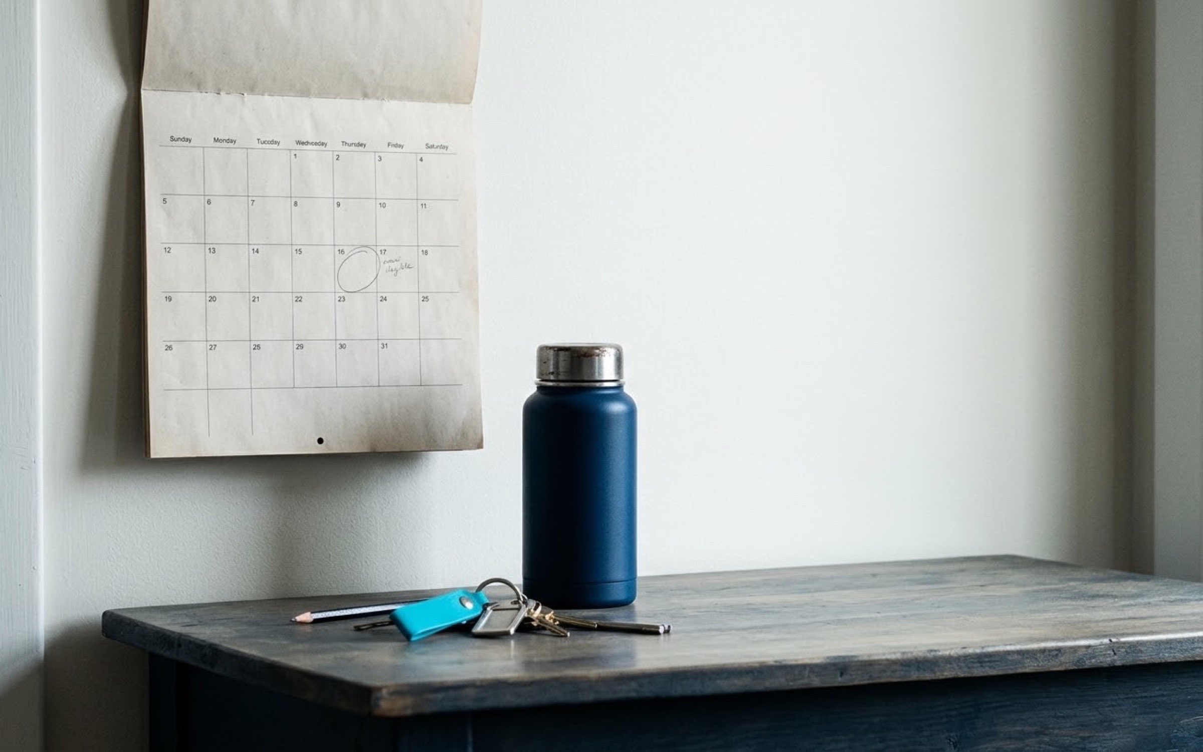 Calendario de pared de papel con una fecha marcada a lápiz, junto a un termo de café y unas llaves sobre una mesa de entrada.