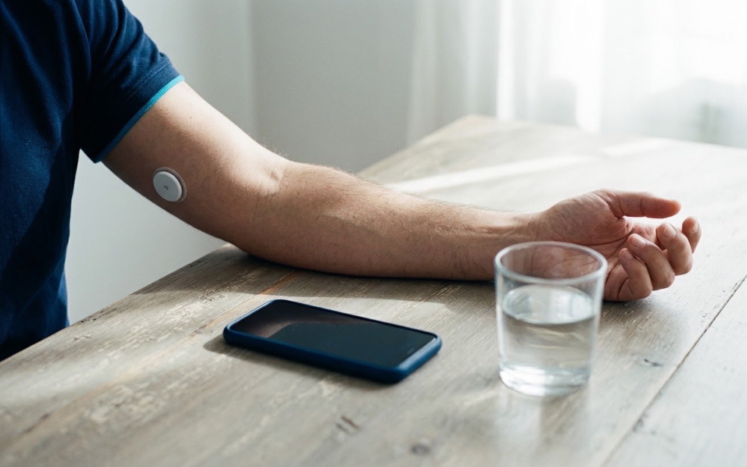 Detalle de un brazo apoyado en una mesa de madera, con un pequeño sensor circular blanco sobre la piel, junto a un teléfono con la pantalla apagada y un vaso de agua.