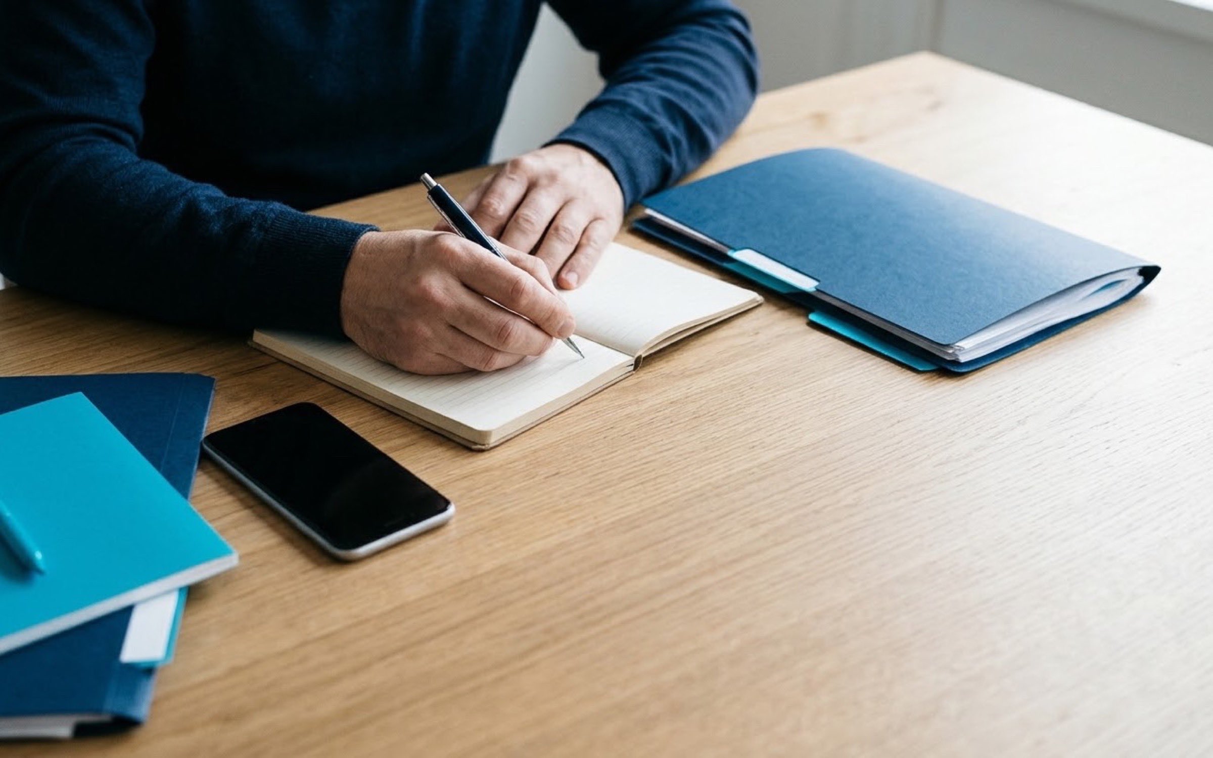 Manos escribiendo en una libreta junto a un celular y una carpeta de papeles ordenados sobre una mesa de madera.