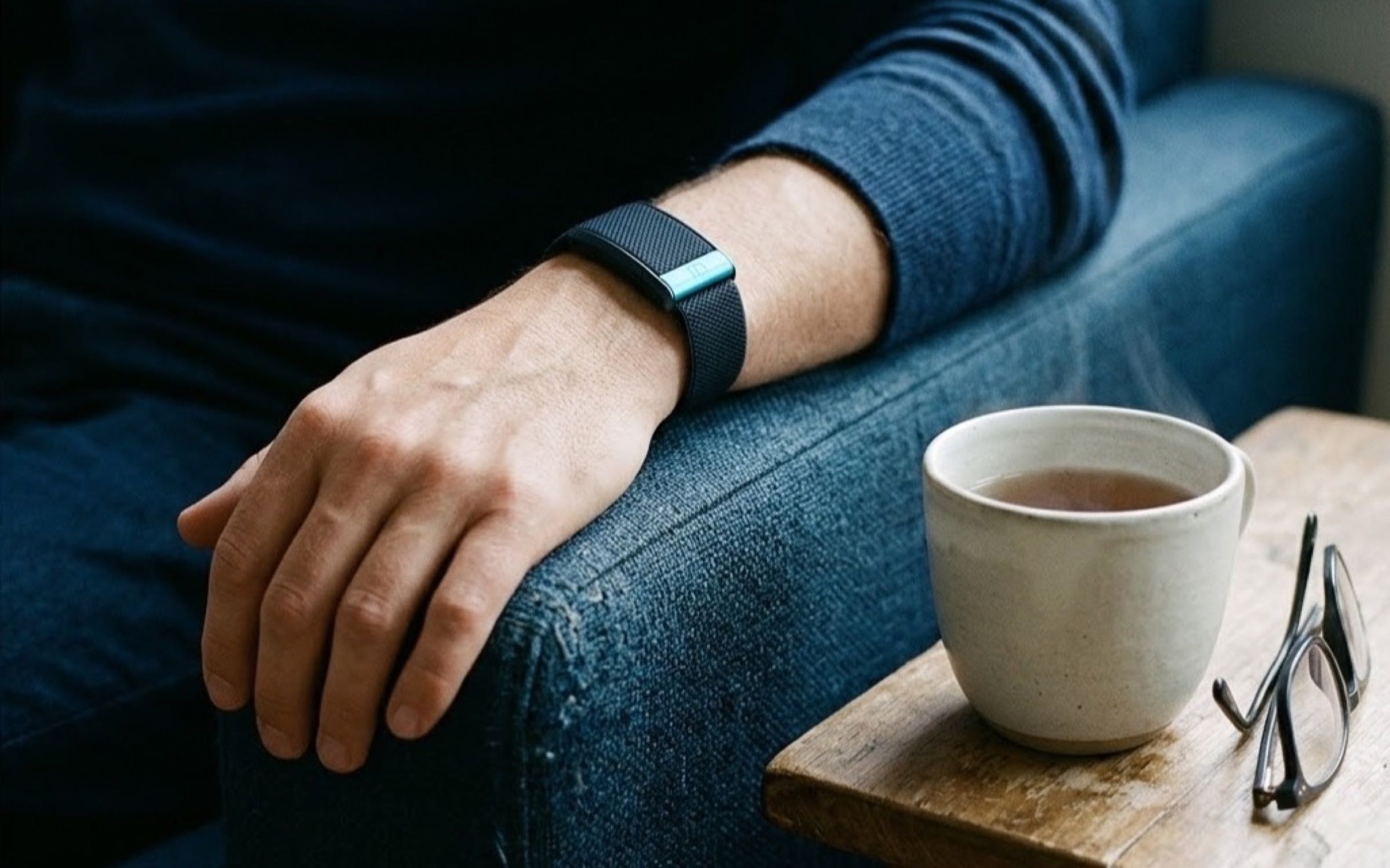 Brazo de una persona con una manilla deportiva apoyado en el brazo de un sillón, junto a una taza de té y unas gafas de lectura sobre una mesa de madera.